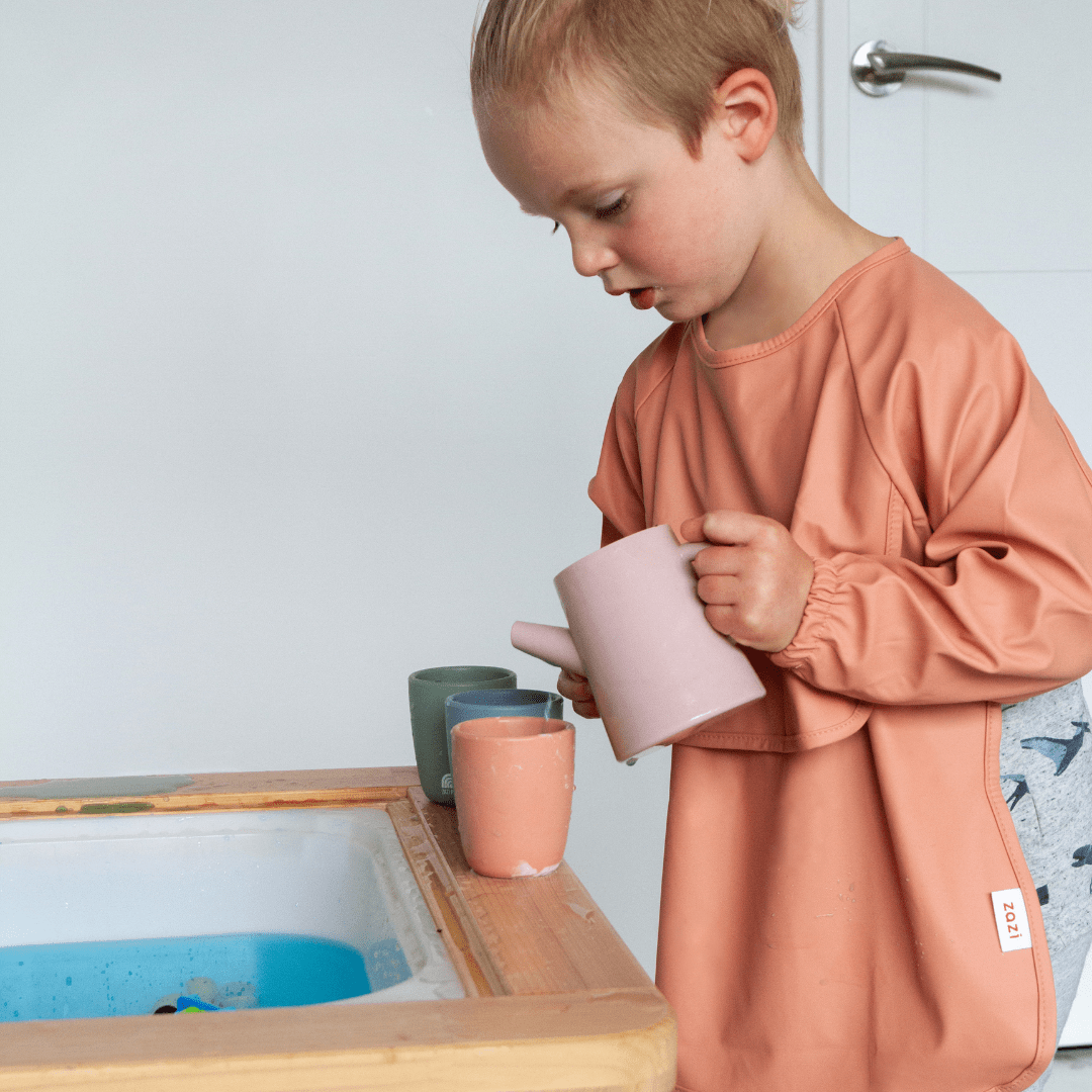 A young boy is standing in front of a table with a Zazi Recycled Full-Sleeved Bib (Multiple Variants).