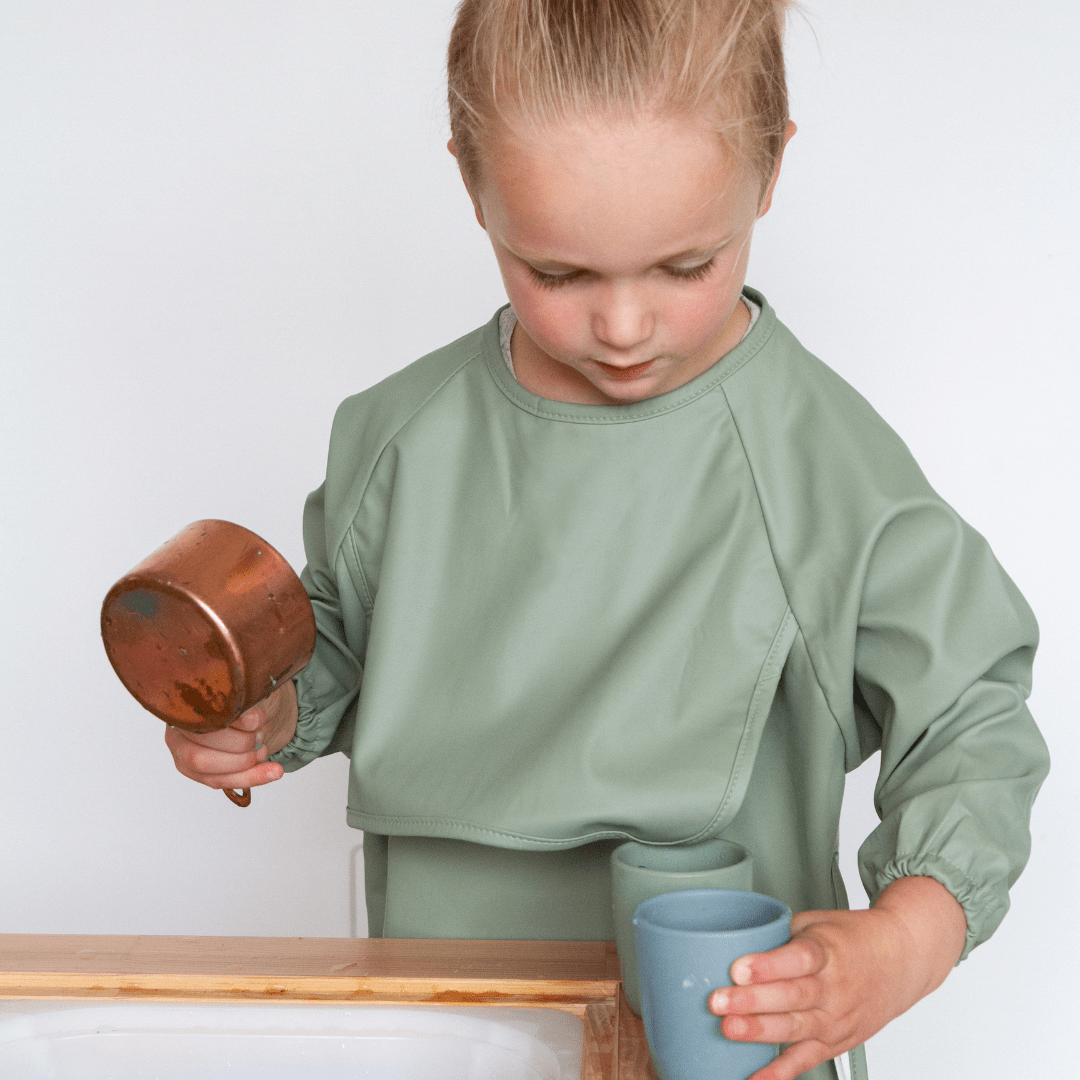 A young child in a green smock is holding a copper cup and a blue cup, standing near a wooden surface, proudly wearing Zazi Recycled Full-Sleeved Bibs that are both waterproof and stain-resistant.