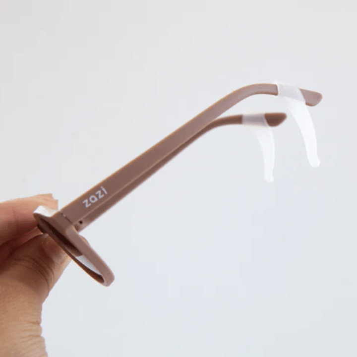 A hand holds a pair of brown eyeglasses with the brand name "Zazi" on the temple. The glasses, designed for a low nose bridge, have clear nose pads.