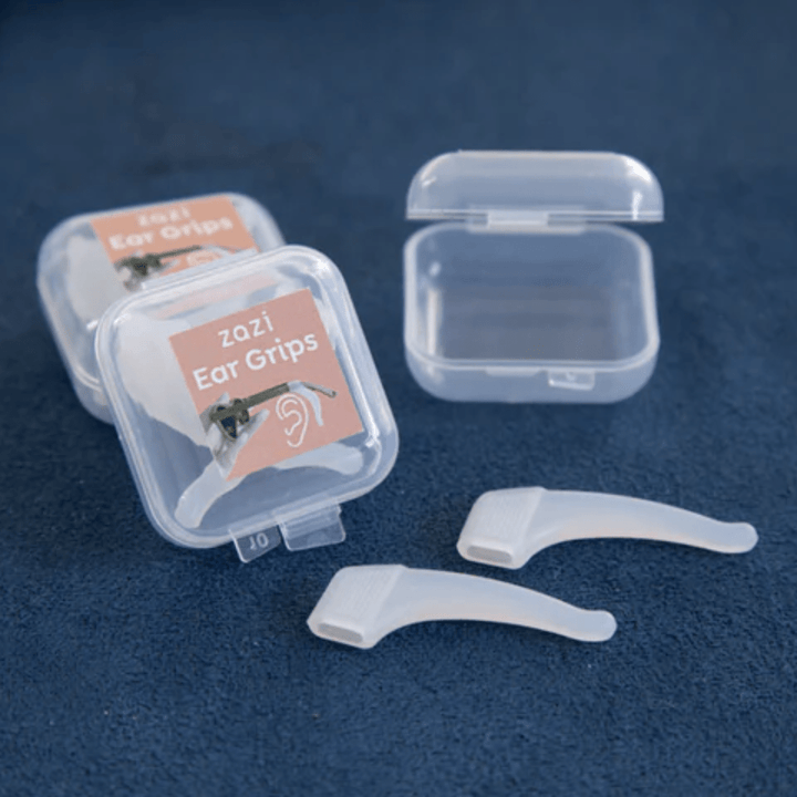 Plastic containers labeled "Zazi Ear Grips" with two white ear grips displayed on a blue surface. One container is open and empty, ideal for holding your Zazi shades or accommodating frames designed for a low nose bridge.