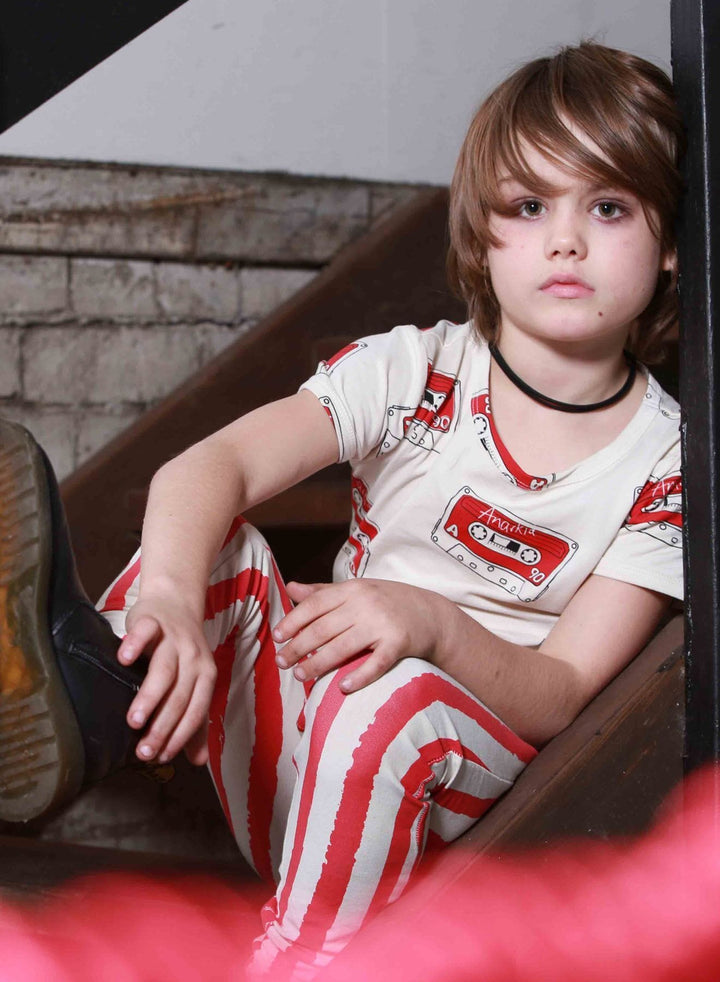 A child with shoulder-length hair sits on a wooden staircase, wearing a white shirt with red cassette tape prints and matching Anarkid Organic Cotton Lollipop Grunge Stripe Leggings - LUCKY LASTS - 0-3 MONTHS & 3 YEARS by Anarkid, one leg bent and resting on a step.