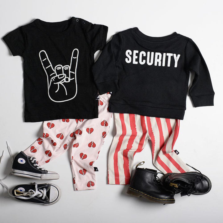Children's clothing set featuring a black T-shirt with a rock hand sign, black sweatshirt with "SECURITY," unisex kids pants including pink heart-print and red-striped designs, Anarkid Organic Cotton Lollipop Grunge Stripe Leggings - LUCKY LASTS - 0-3 MONTHS & 3 YEARS by Anarkid, complemented by black sneakers and boots—all on a white background.