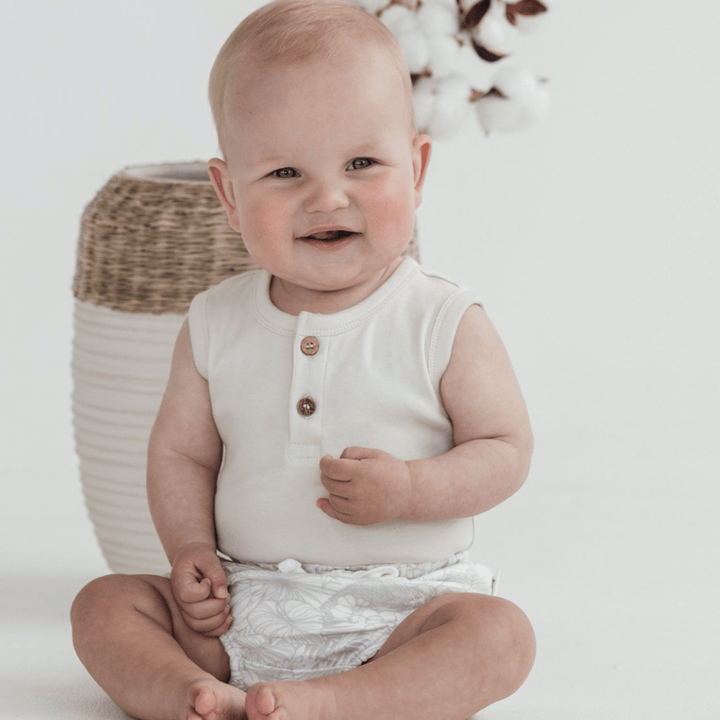 A baby sits on the floor wearing a sleeveless top and Aster & Oak's Aster & Oak Organic Animal Bloomers - LUCKY LASTS - 3-6 MONTHS & 6-12 MONTHS ONLY, smiling. There is a woven basket with cotton plants in the background.