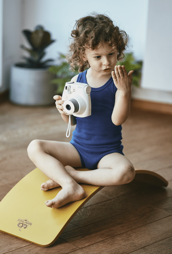 A child with curly hair, dressed in a blue tank top, sits cross-legged on a yellow Kinderfeets Balance Board by Kinderfeets indoors. Holding a white instant camera and looking at their hand, this scene exemplifies open-ended play, fostering creativity and supporting child development.