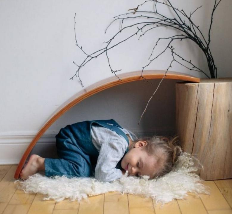 A child sleeps on a fluffy rug under a curved wooden structure, reminiscent of the Kinderfeets Balance Board by Kinderfeets, next to a wooden planter with branches. This serene scene fosters both rest and open-ended play, essential for child development.