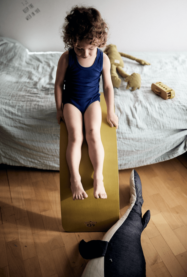 A child with curly hair in a blue outfit sits on a yellow slide indoors, surrounded by toys and a bed in the background, with a Kinderfeets Balance Board from Kinderfeets nearby. The scene is perfect for open-ended play that fosters child development.