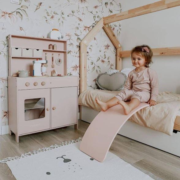 A young child sits on a small bed in a room with floral wallpaper. Next to the bed are a pink play kitchen and a Kinderfeets Balance Board by Kinderfeets for open-ended play. On the floor, there's a white rug with simple designs, fostering child development in a nurturing environment.