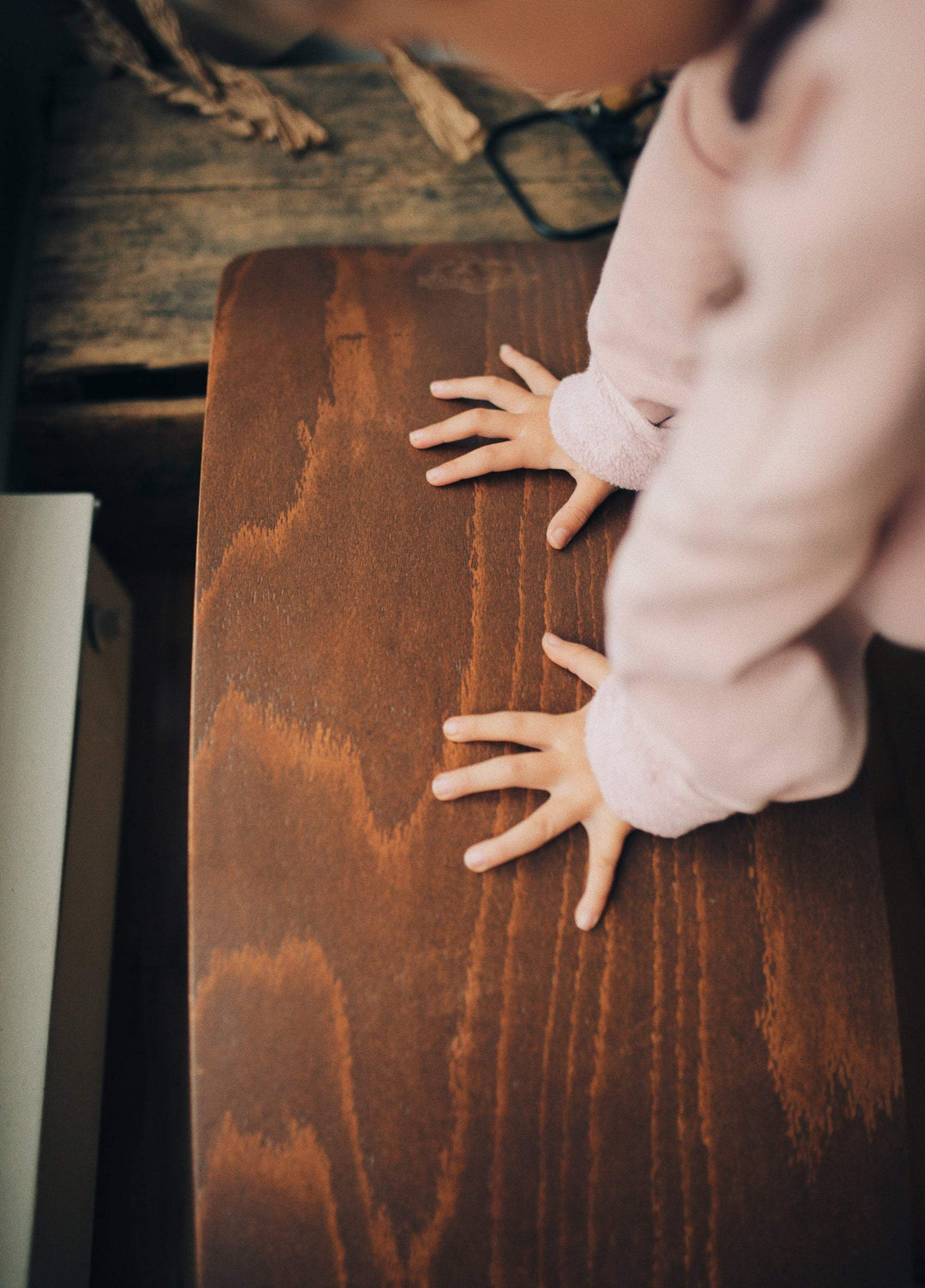 A child wearing a pink sleeve shirt places their hands on a brown wooden surface, engaging in open-ended play with the Kinderfeets Balance Board by Kinderfeets. Glasses are visible in the background.