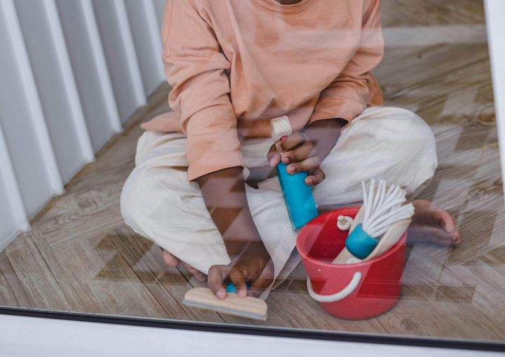 A child sits on the floor, fully engrossed in imitative play, holding a cleaning brush and spray bottle from the Plan Toys Cleaning Set - LUCKY LAST, crafted by Plan Toys from sustainable rubber wood, with a red bucket nearby.