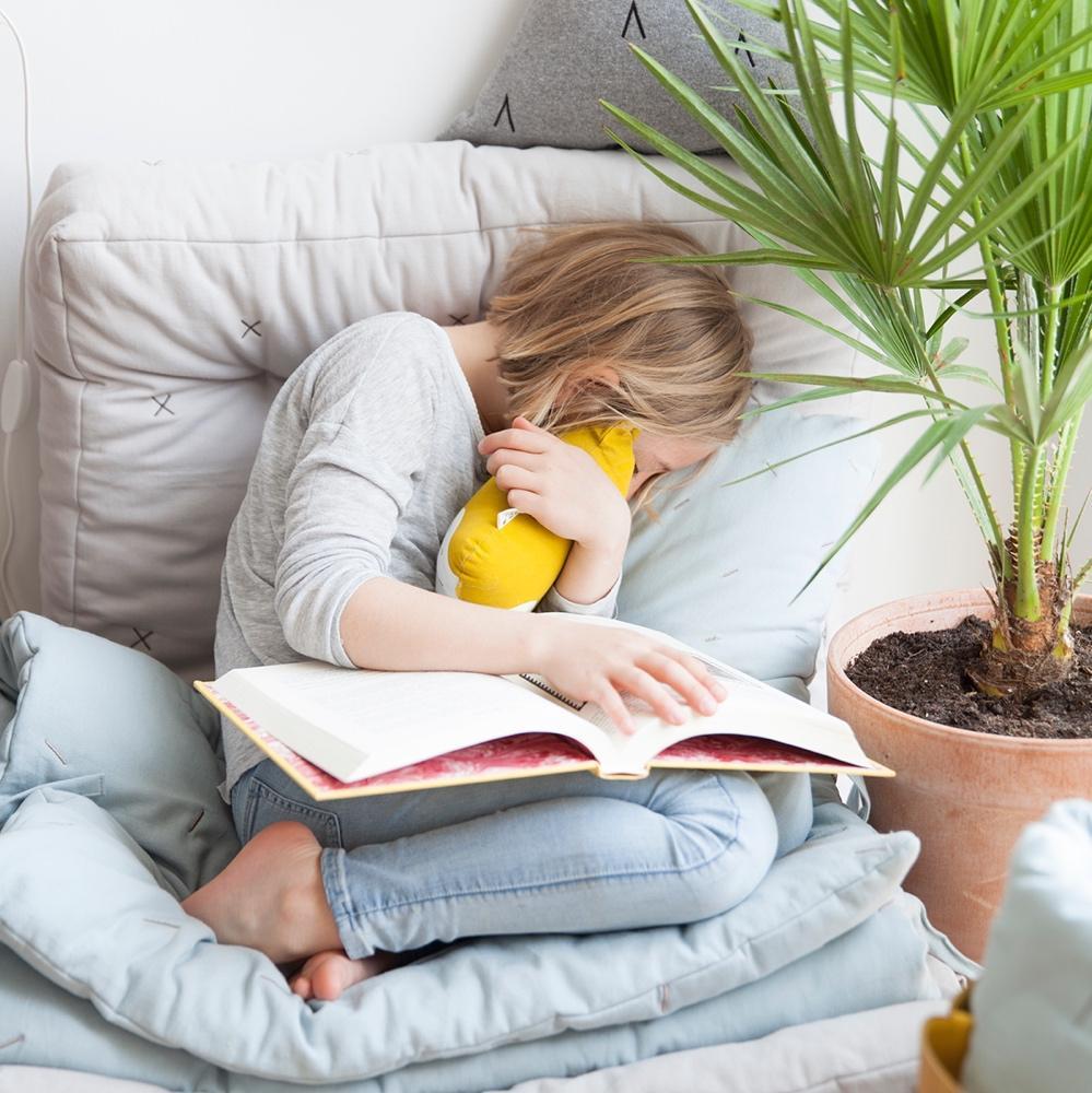 A girl is reading a book while sitting on a Fabelab Organic Cotton Animal Cushion - LUCKY LAST - BEAR (HONEY).