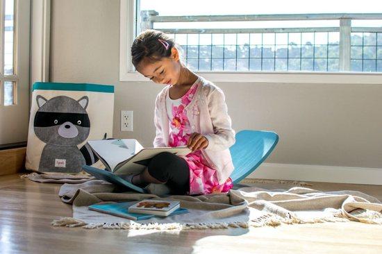 A young girl in a pink dress and white cardigan sits on the floor, engrossed in a book. Behind her is a stuffed raccoon toy and a Kinderfeets Balance Board (Multiple Variants), fostering child development through open-ended play.