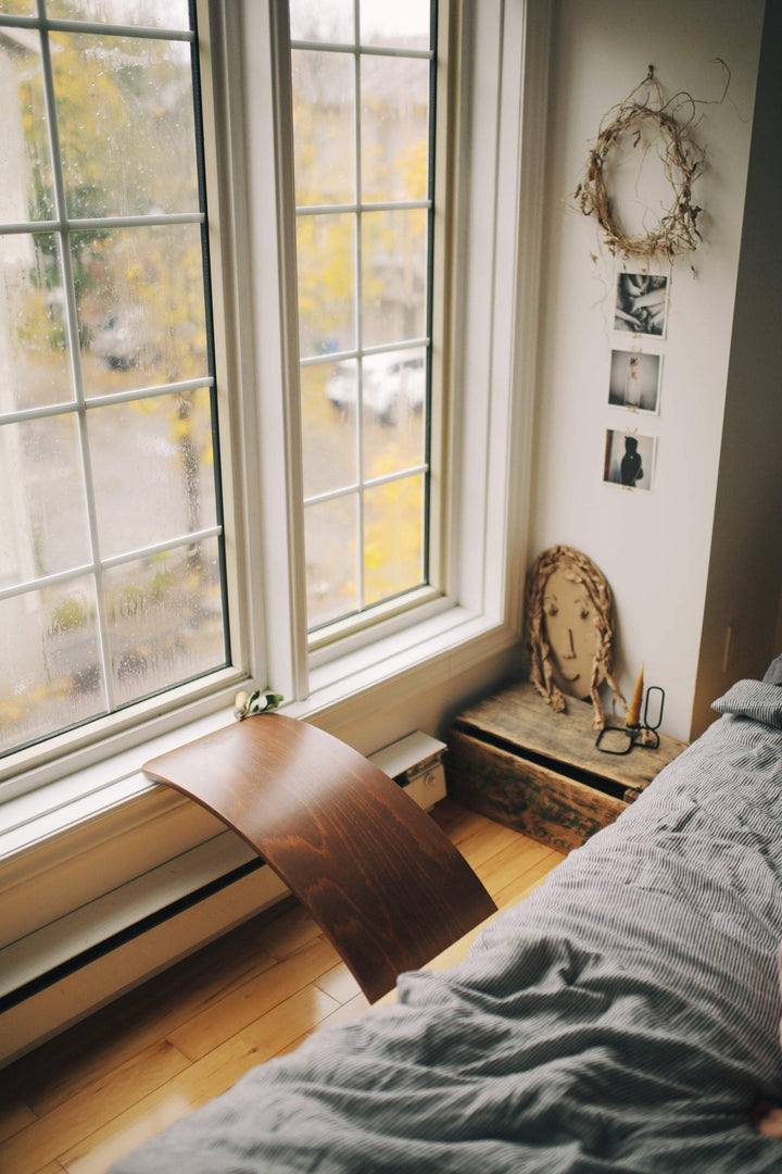 A cozy bedroom with a large window, a wooden side table pressed against the wall, photos adorning the walls, a small shelf holding an old book and a pair of eyeglasses, and a bed dressed in gray bedding. On the floor lies a Kinderfeets Balance Board by Kinderfeets, inviting open-ended play that is perfect for child development.