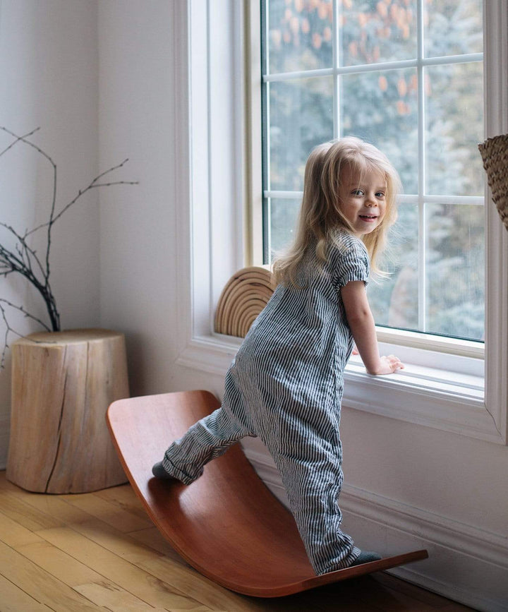 A young child in striped pajamas stands on a Kinderfeets Balance Board by a large window, gazing outside. Embracing open-ended play, the scene is complemented by a tree stump stool and bare branches, creating an environment that fosters child development.