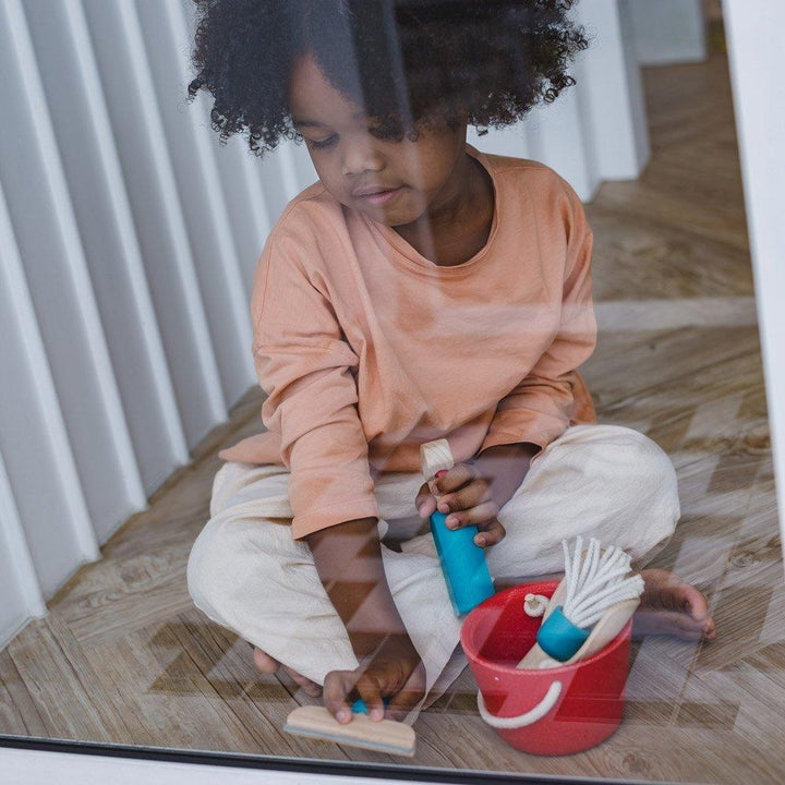 A child in a peach shirt and white pants is sitting on a wooden floor, engaging in imitative play with their Plan Toys Cleaning Set - LUCKY LAST, featuring a red bucket and a mop made from sustainable rubber wood by Plan Toys.