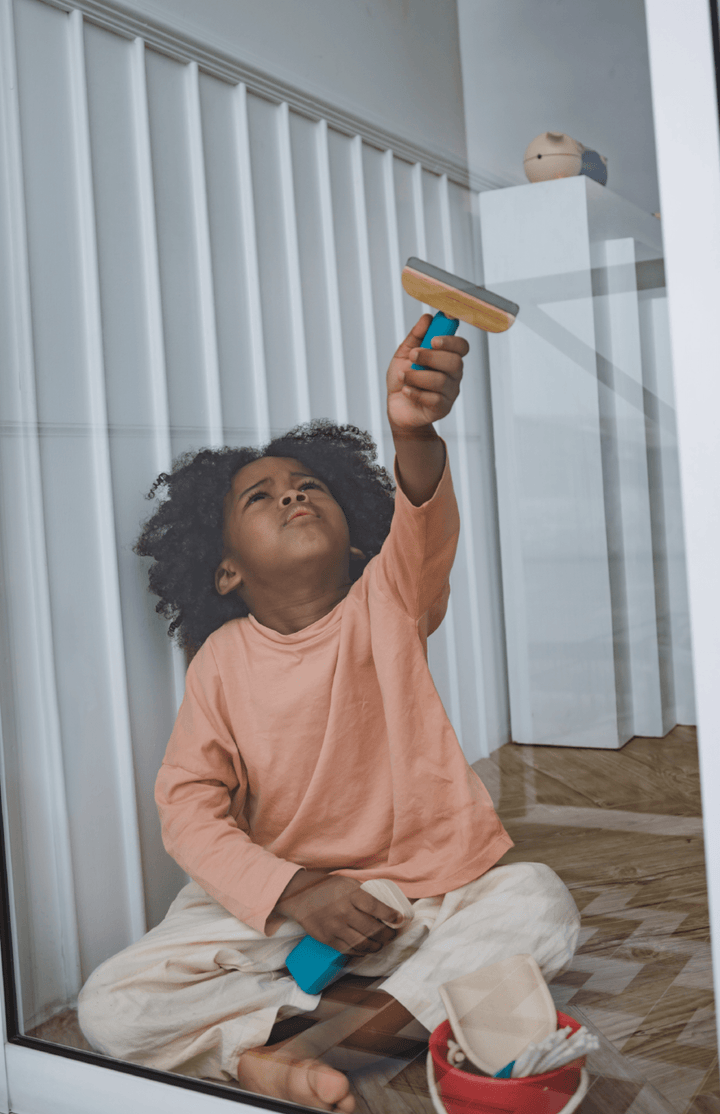 A child sits focused on the floor, holding a toy squeegee from the Plan Toys Cleaning Set - LUCKY LAST. Engaged in imitative play, they press it against a glass surface, captivated by their task. Designed by Plan Toys and crafted from sustainable rubber wood, this toy inspires eco-friendly fun.