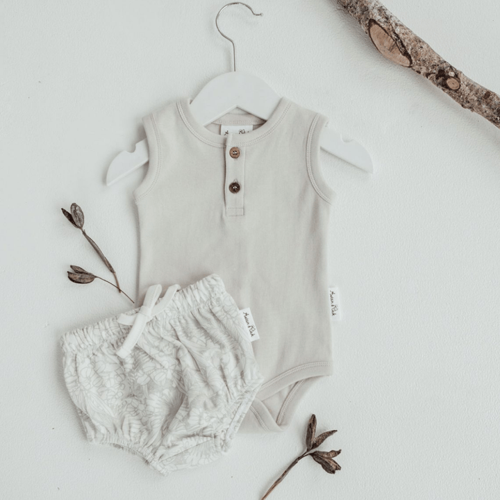 A beige baby bodysuit with buttons and white floral shorts on a hanger, displayed against a white background with decorative twigs, featuring Aster & Oak Organic Animal Bloomers - LUCKY LASTS - 3-6 MONTHS & 6-12 MONTHS ONLY for added comfort.